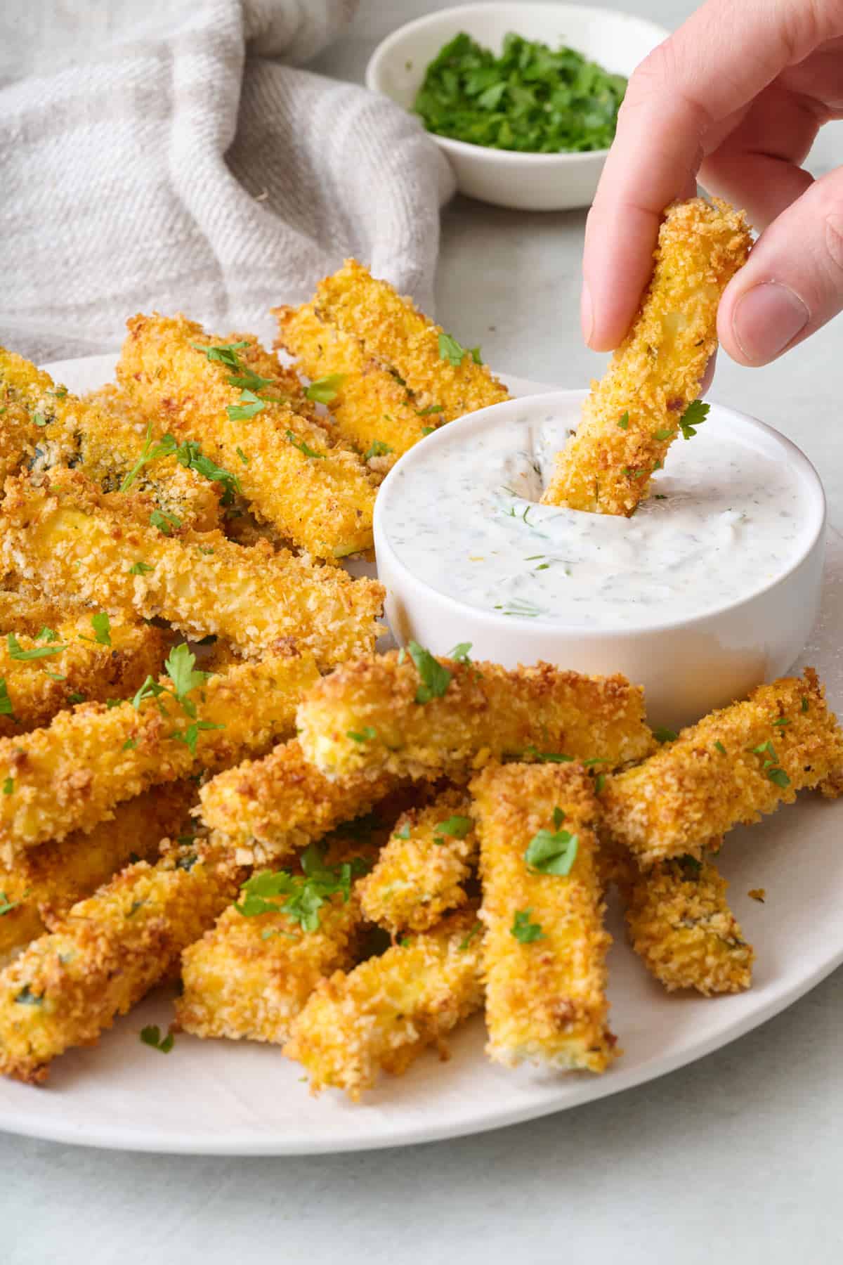 A person dipping a baked zucchini fry into a bowl of dip set on a platter with baked zucchini fries.