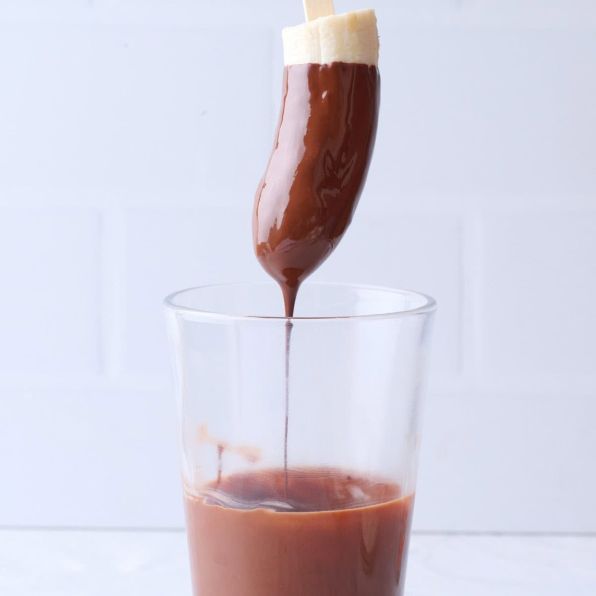 Banana dipped into a cup of melted chocolate being lifted up with chocolate drizzling back into cup.