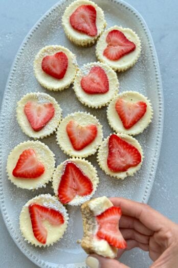 Cottage Cheesecake cups on a platter topped with strawberries with hand holding one of them bitten out of