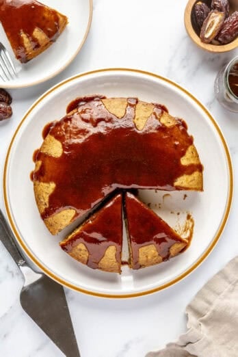 Date cake topped with date syrup on a round cake plate with a few slice cut away.