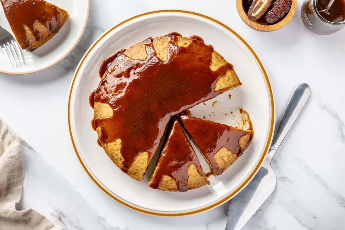 Date cake topped with date syrup on a plate with a few slices cut away.
