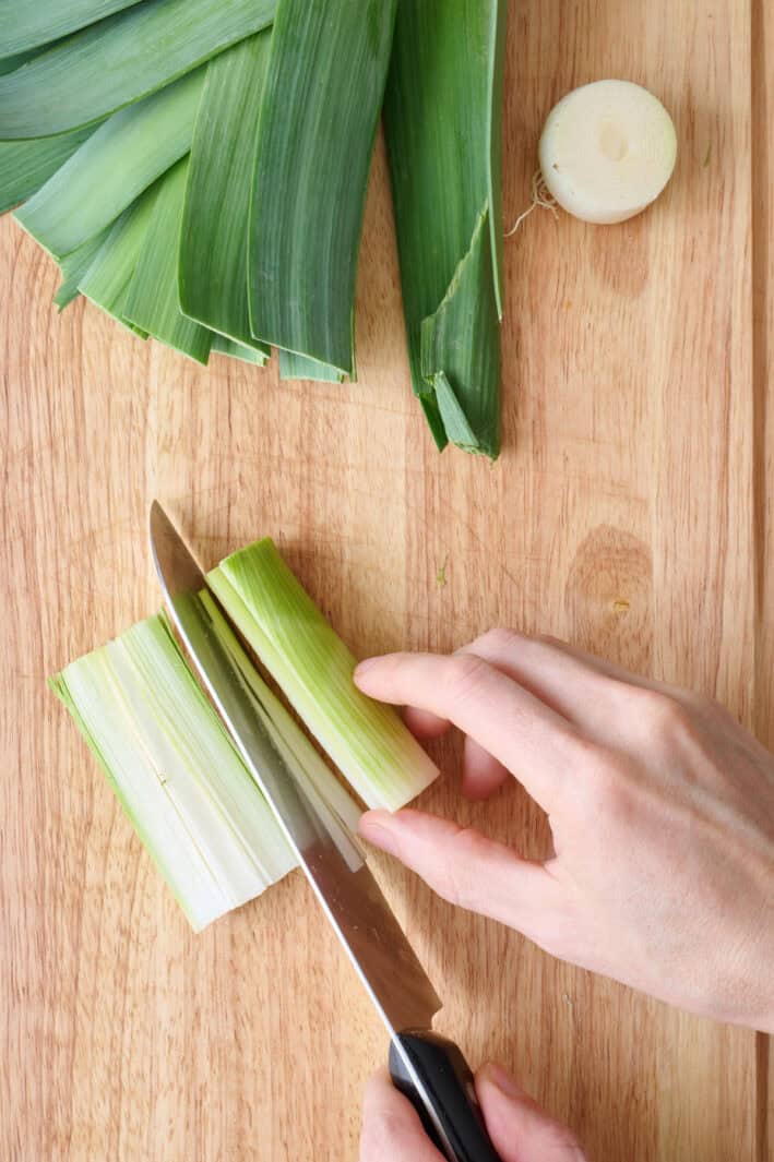 Tutorial for how to cut and wash leeks with a trimmed stalk on cutting board being cut in half.