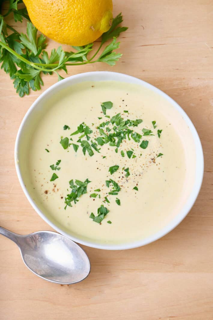 Hollandaise sauce in a serving bowl, garnished with a small amount of fresh parsley and pepper, spoon and lemon nearby.