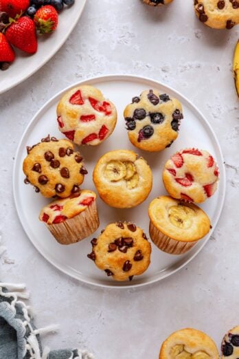 Pancake muffins on a round plate with different add ins including strawberry, blueberries, chocolate chips, and bananas.