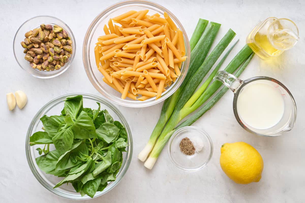 Ingredients for recipe in individual dishes: pistachios, garlic, basil, penne, green onions, oil, salt and pepper, milk, and lemon.