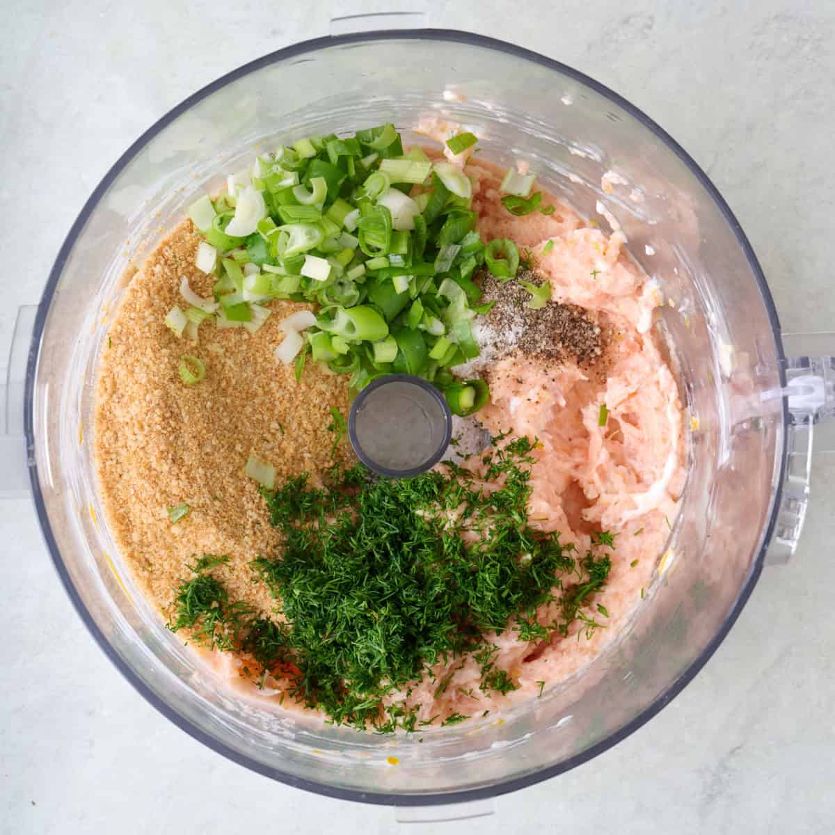 Salmon mixture, dill, scallions, and breadcrumbs in the bowl of a food processor.