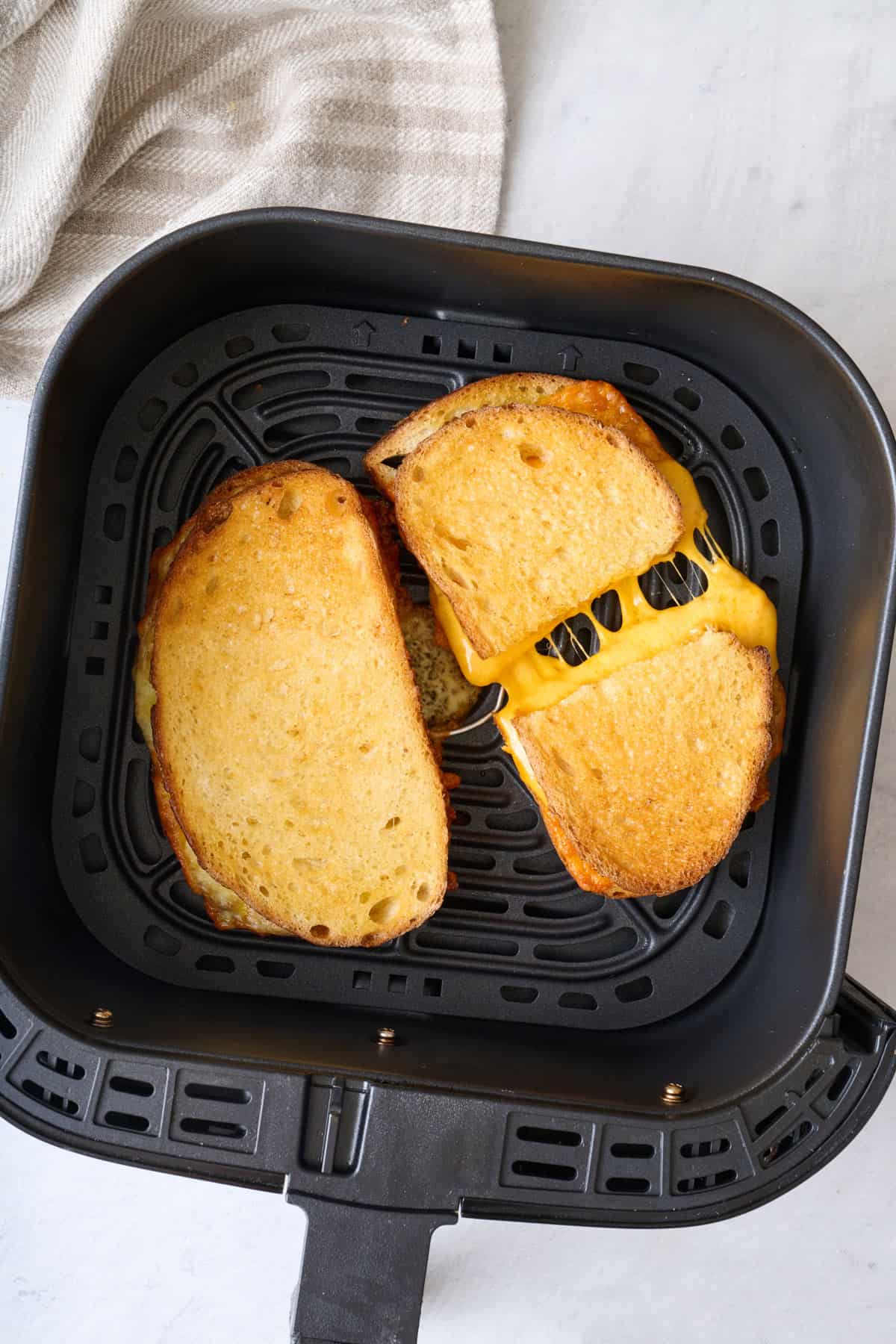 2 grilled cheese sandwiches in an air fryer basket, one cut and slightly pulled apart to show melty cheese.