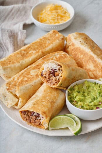 Ground beef burritos on a plate with guacamole and lime nearby.