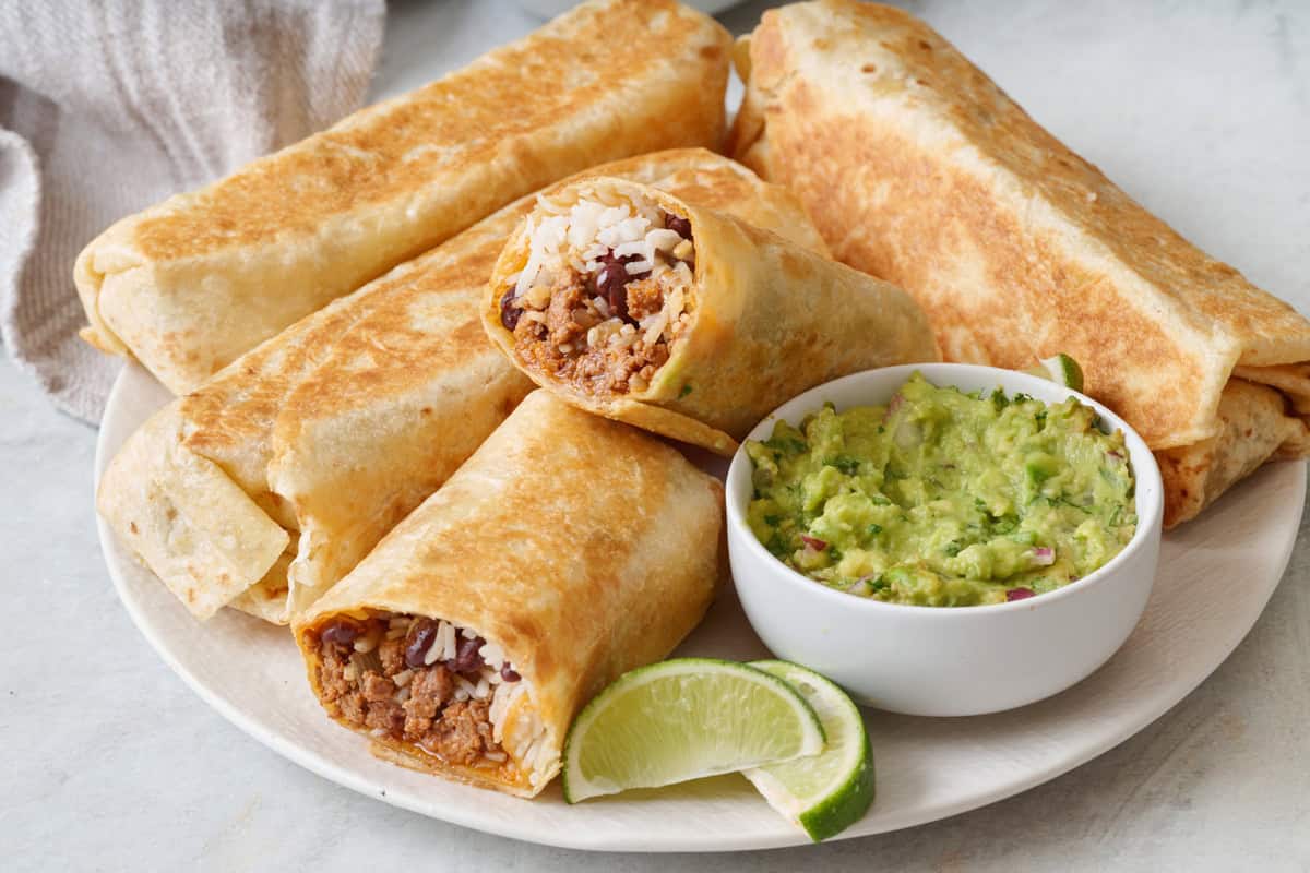 Ground beef burritos on a plate with guacamole and lime nearby.