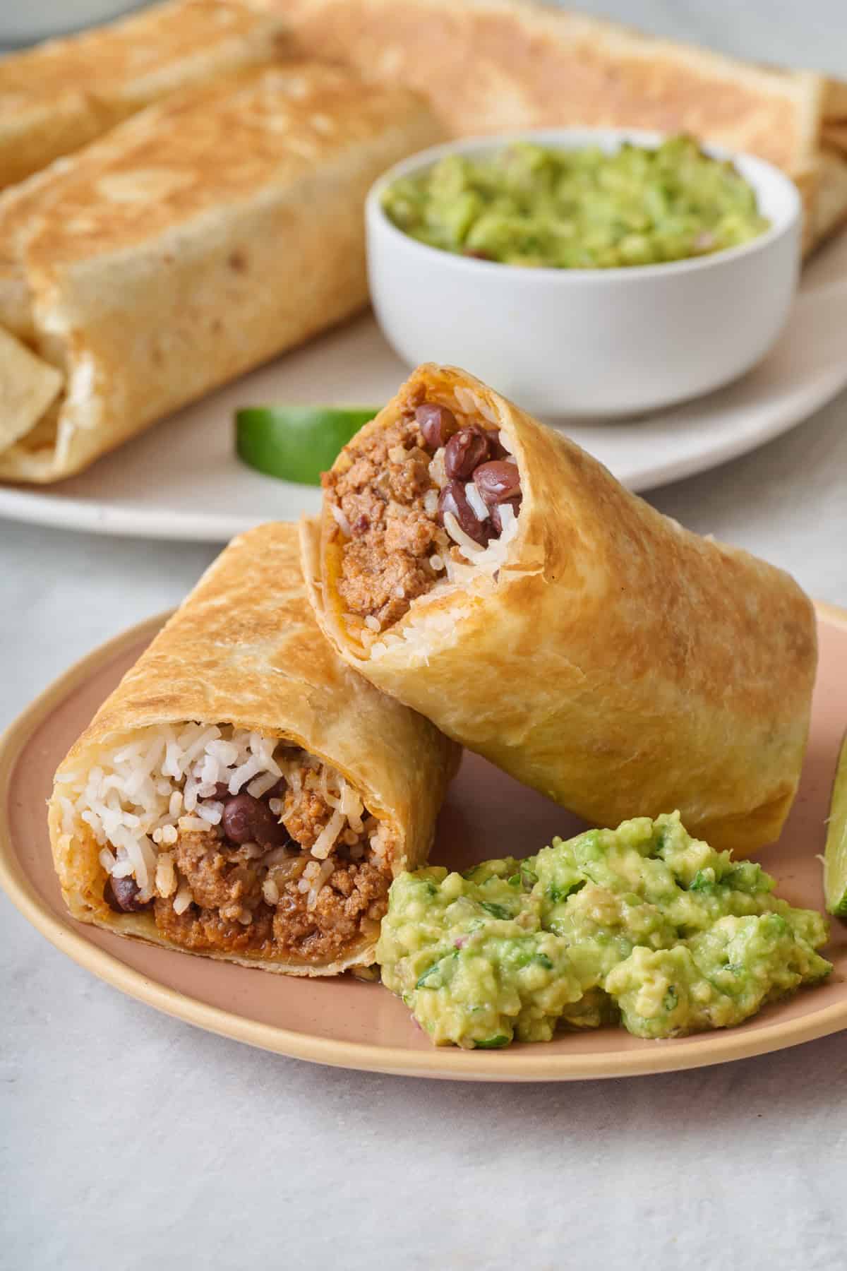 Ground beef burrito on a plate with a side of guacamole and more burritos nearby.