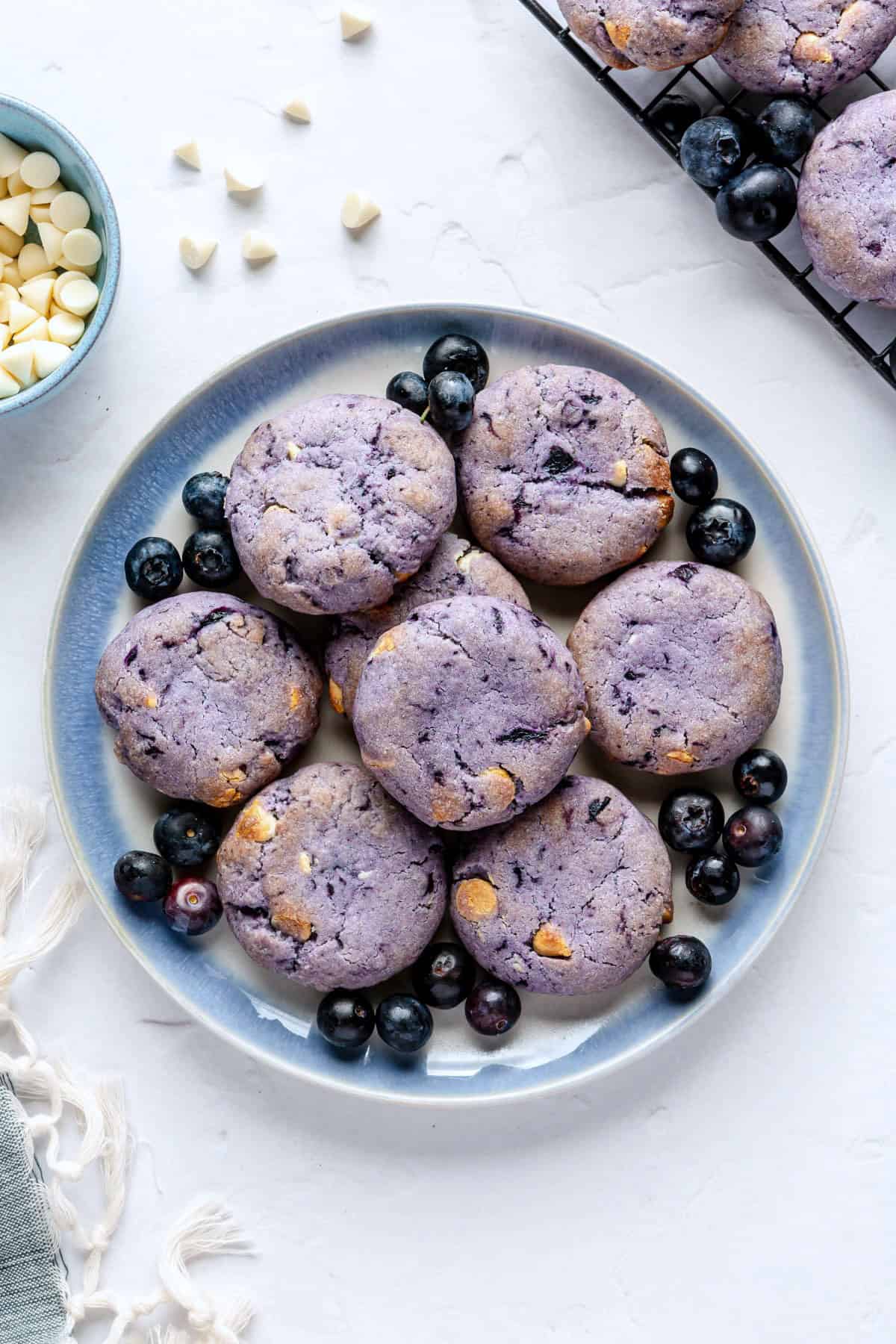 A few Blueberry cookies on a small round plate with small dish of white chocolate chips and extra blueberries around, wire rack of more cookies nearby.