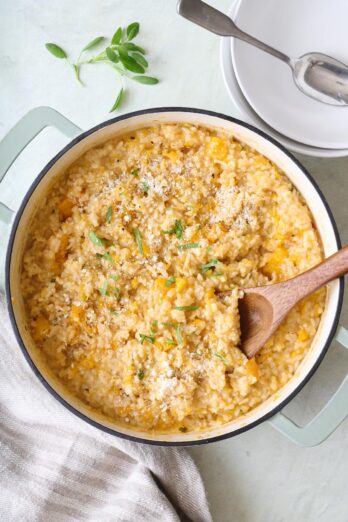 Butternut squash risotto in a pot garnished with parmesan and sage with a wooden serving spoon dipped in pot.