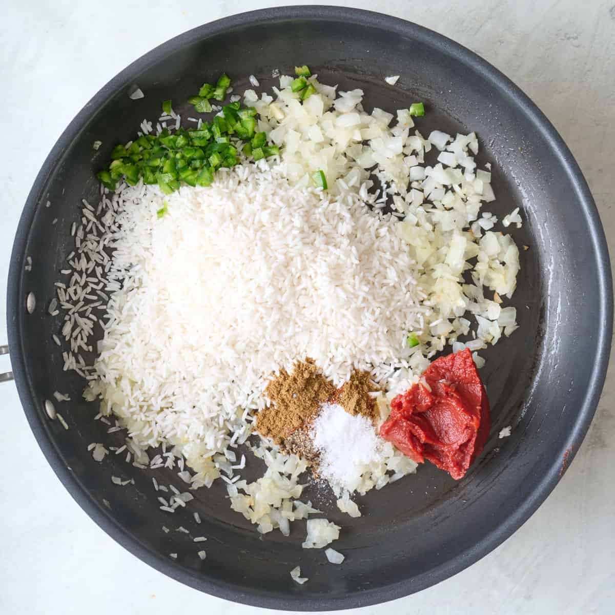 Chopped veggies, spices, tomato paste, and uncooked rice in a skillet before mixing.