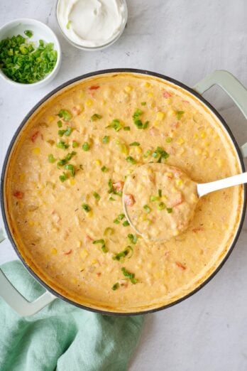 Pot of corn chowder with diced green onions sprinkled on top and a ladle dipped inside lifting some up. Small dishes of extra green onions and yogurt nearby.