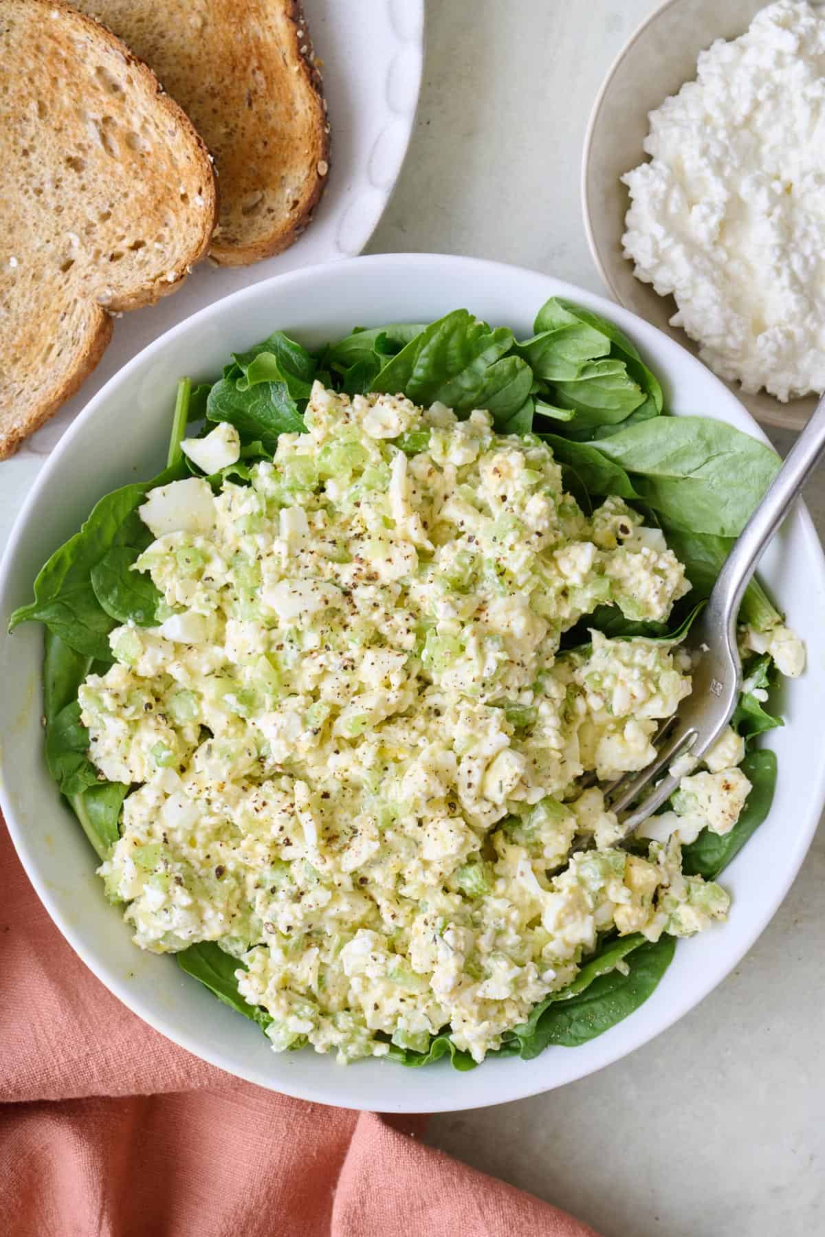 Egg salad in a bowl on top of fresh greens with a fork dipped in, with toast and cottage cheese nearby.