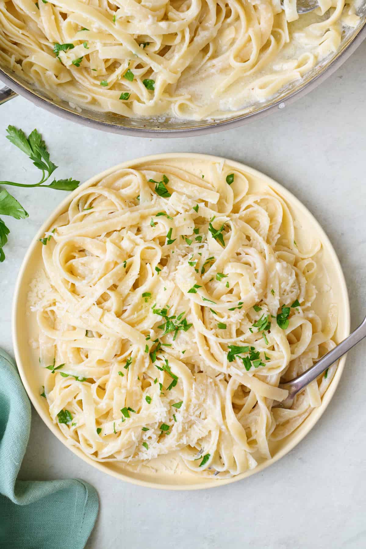 A serving of Fettuccine alfredo on a plate with a fork dipped in, garnished with fresh parsley and parmesan cheese.