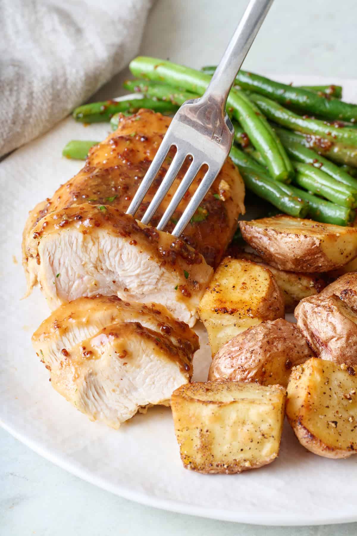 Fork lifting up a slice of hone mustard chicken breasts from a plate, served with green beans and potatoes.