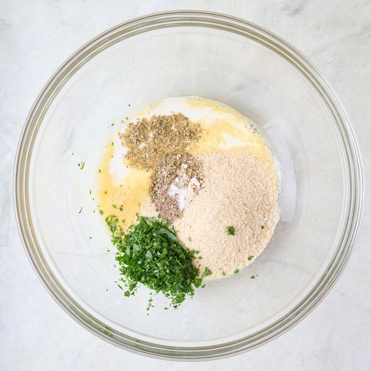 Spices, herbs, milk, egg, and breadcrumbs in a large glass mixing bowl.