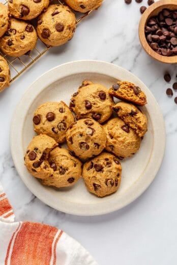 Pumpkin chocolate chip cookies on a plate with wire rack of more cookies and bowl of chocolate chips nearby.