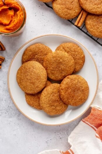 A few cookies on a round plate with wire rack of more cookies nearby, cinnamon sticks and small jar of pumpkin puree nearby.