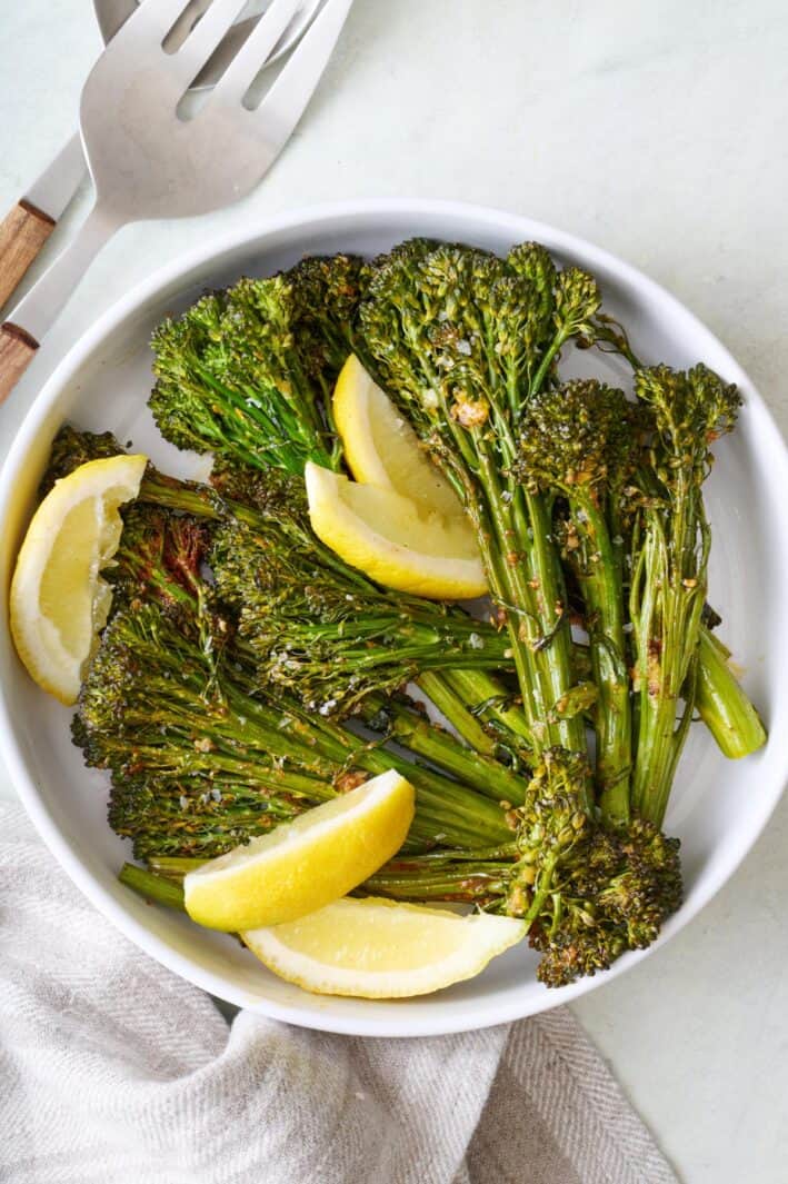 Bowl of seasoned roasted broccolini, garnished with lemon wedges.