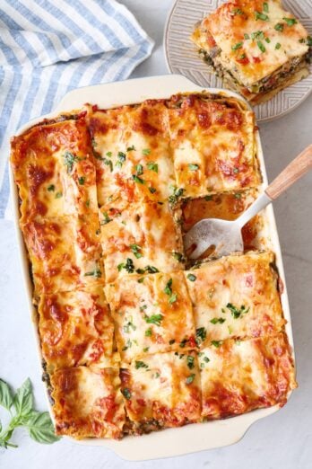 Vegetable lasagna in baking dish after cutting into 12 pieces with one slice removed to a plate on the side, and serving utensil in dish.