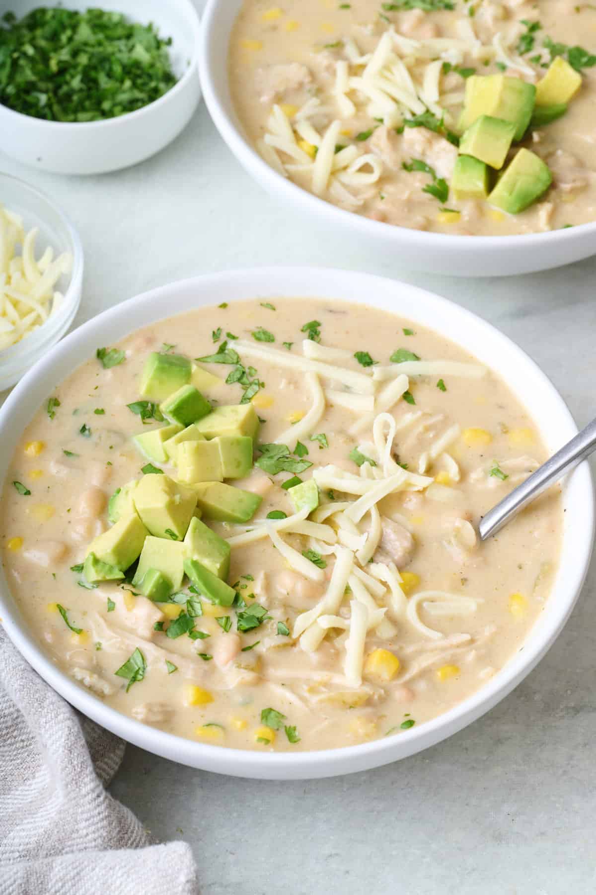 Two bowls of creamy white chicken chili with shredded cheese diced, avocado on top.