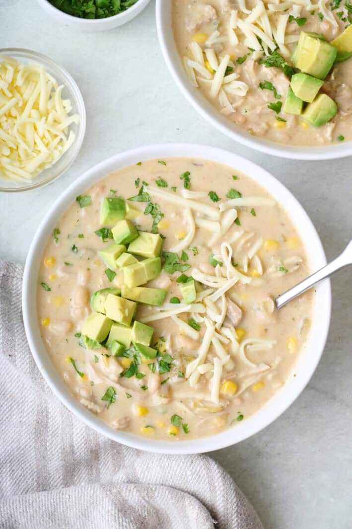 Two bowls of creamy white chicken chili with shredded cheese diced, avocado on top.