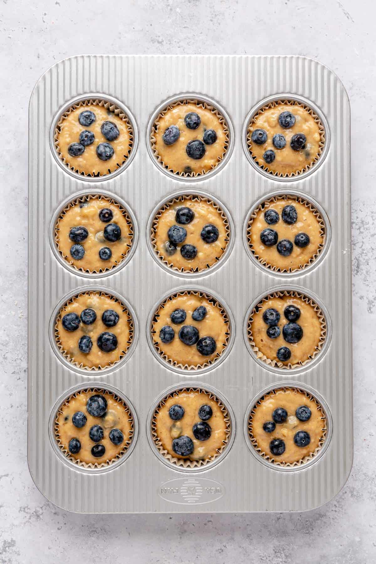 Muffin batter added to a 12 cup muffin tin with a paper liners and extra blueberries added on top.