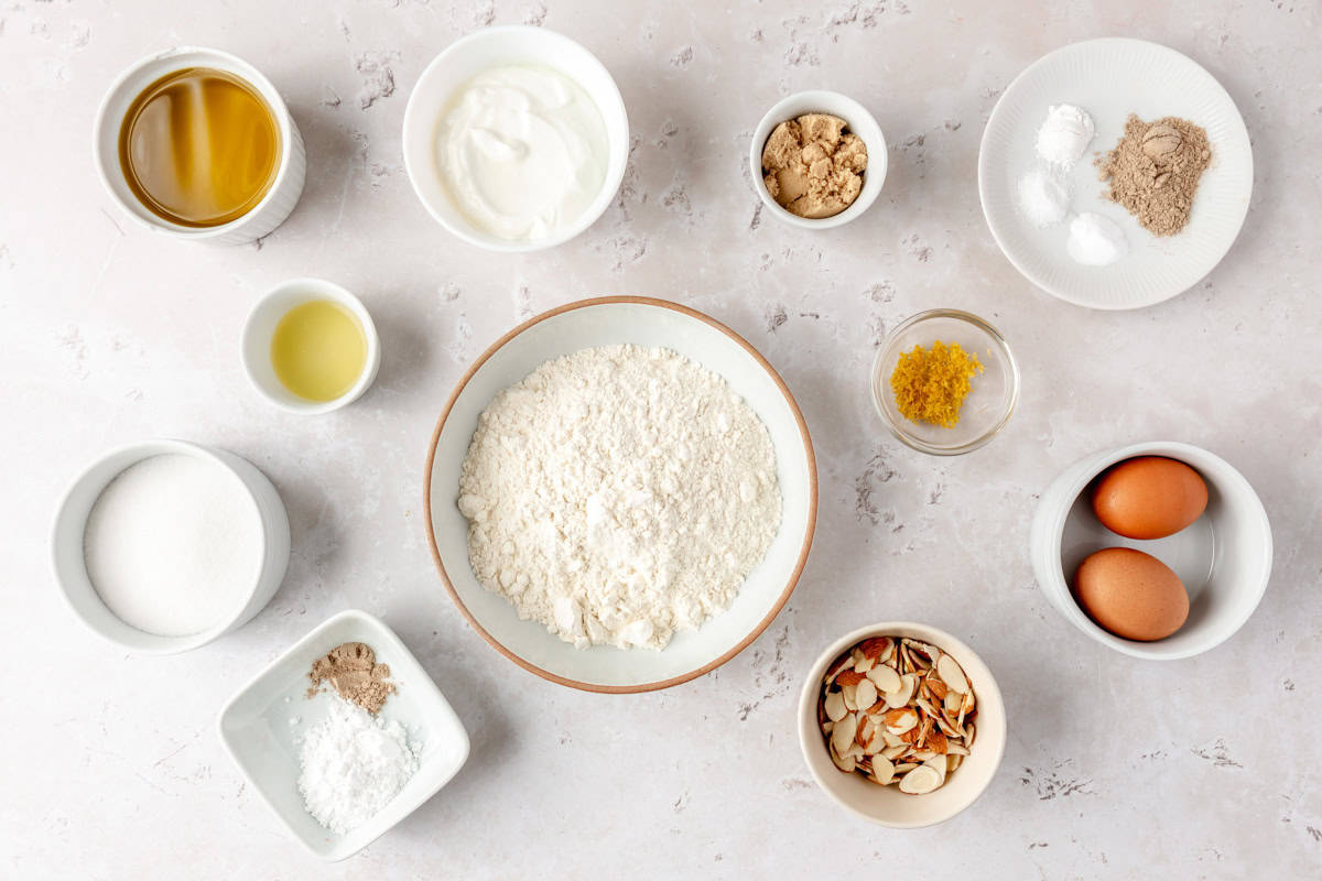 Ingredients for recipe in individual bowls: olive oil, lemon juice, sugar, powder sugar and cardamom, greek yogurt, brown sugar, lemon zest, sliced almonds, eggs, baking powder, baking soda, ground cardamom, and salt.