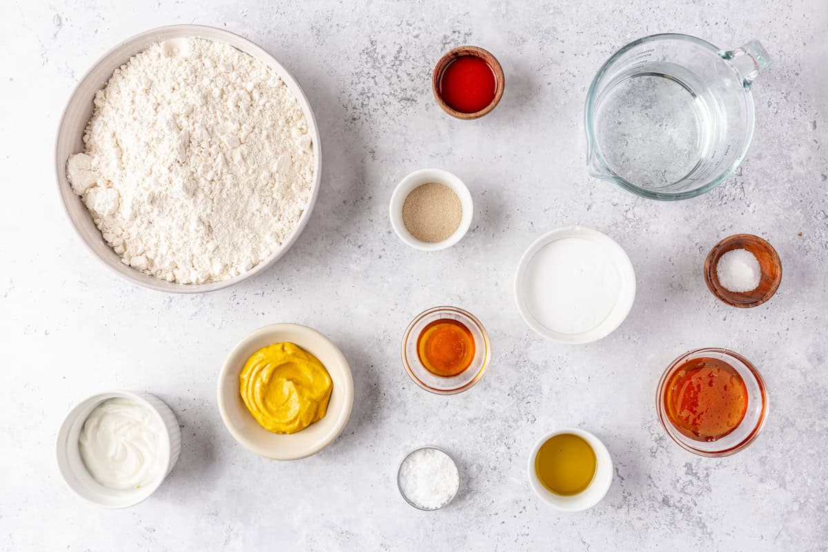 Ingredients for recipe in individual bowls: flour, yeast, olive oil, honey, salt, sea salt, baking soda, plus ingredients for mustard sauce including yellow mustard, honey, greek yogurt, and hot sauce.