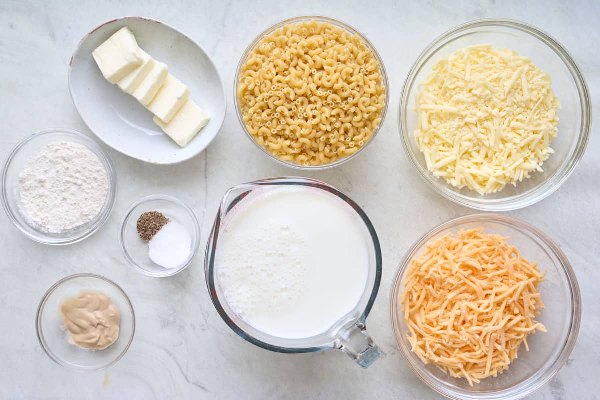 Ingredients for recipe in individual bowls: flour, dijon mustard, butter, salt and pepper, dry elbow macaroni noodles, milk, shredded white cheddar cheese, and shredded sharp cheddar cheese.