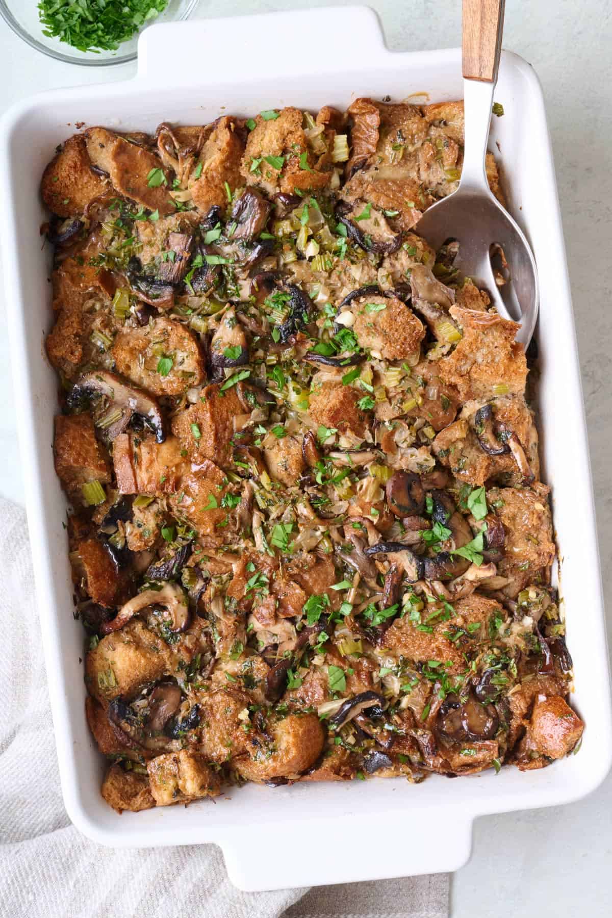 Mushroom stuffing in a baking dish with extra parsley on top and a serving spoon dipped in.