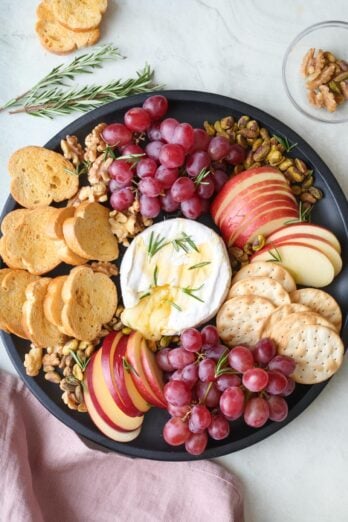 Baked brie platter with cheese cut open and melting out surrounded by apple slices, crackers, grapes, walnuts and pistachios, and toasted bread slices.