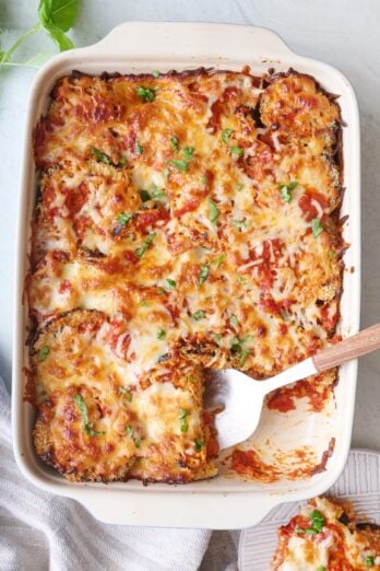 Eggplant parmesan in baking dish with one stack removed to a plate on the side, and serving utensil in dish.