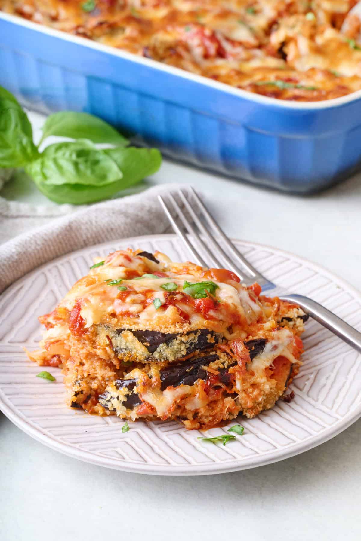 Serving of eggplant parmesan on a small plate showing layers of recipe with baking dish nearby.