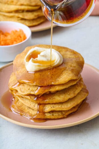 Pouring syrup over a stack of carrot cake pancakes topped with whipped cream cheese.