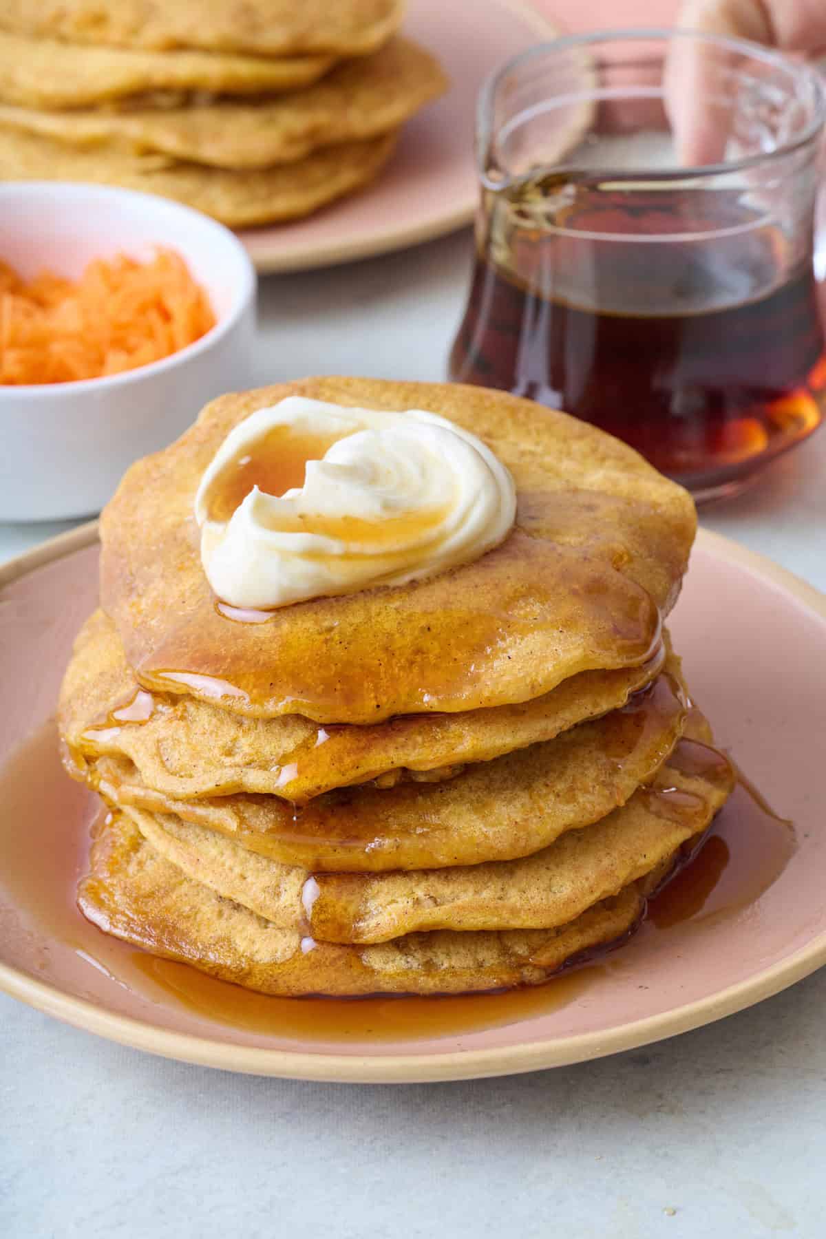 Stack of carrot cake pancakes on a plate topped with sweetened whipped cream cheese with some drizzling down the side, small dish of shredded carrots and syrup nearby.