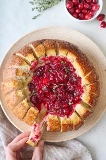 Hand pulling a slice of bread away from bread bowl with baked brie and cranberries.