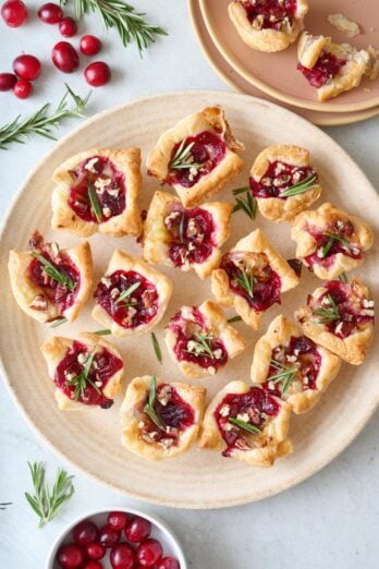 Cranberry brie bites on a platter with fresh rosemary on each, a few fresh cranberries and extra sprigs around. Small plate with a few bites on it nearby.