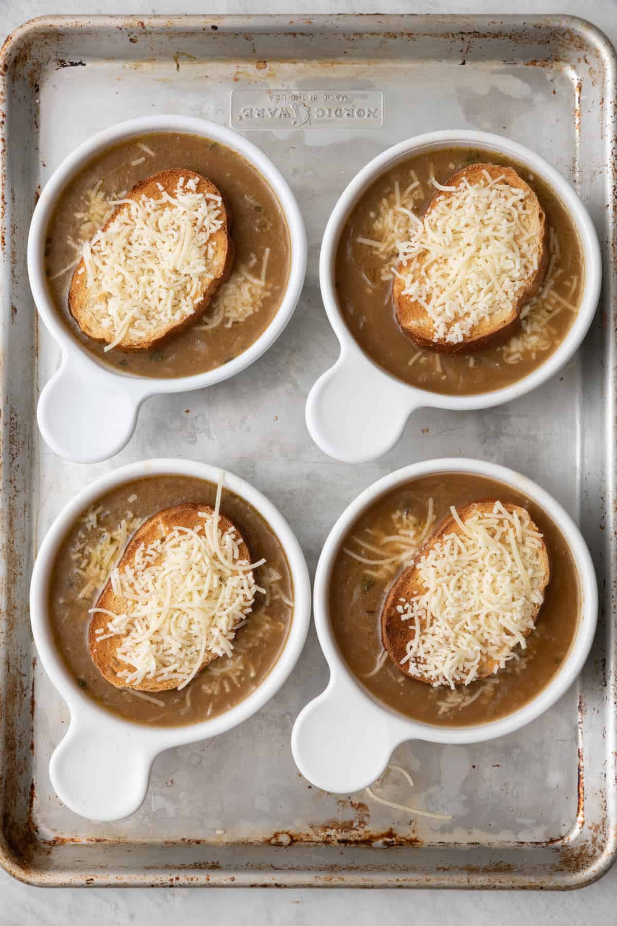 4 bowls on a sheet pan filled with soup, a toasted bread and topped with cheese before broiling.