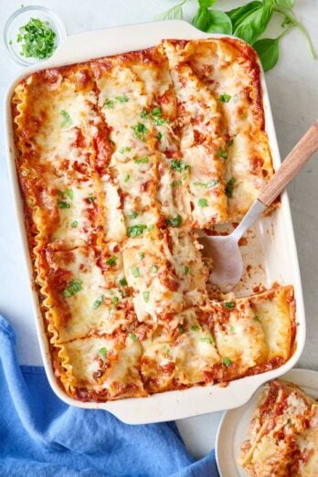 Lasagna in baking dish after cutting into 12 pieces with one slice removed to a plate on the side, and serving utensil in dish