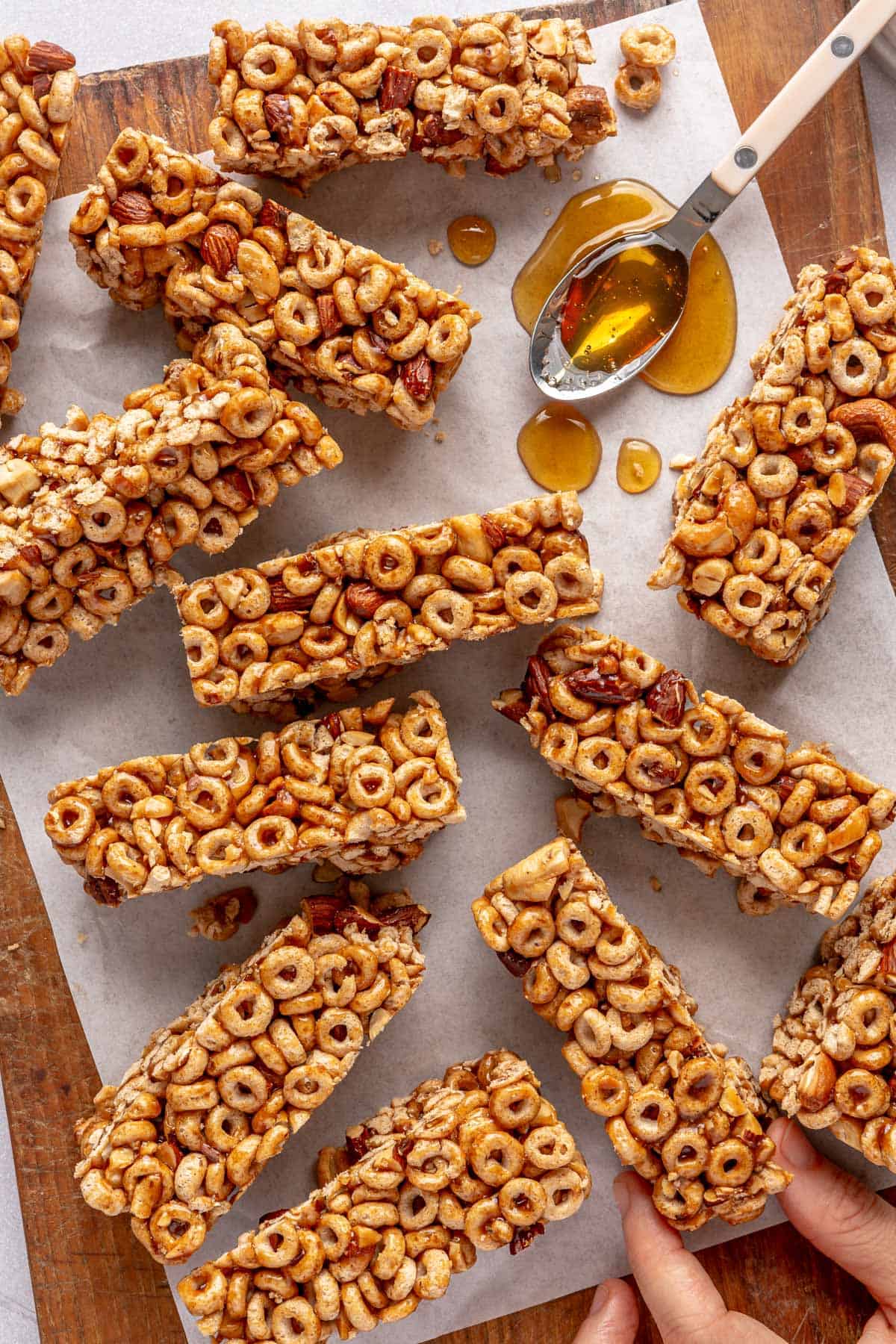 Homemade cereal bars on a cutting board after cut with a spoon of honey resting on parchment paper with a hand grabbing one bar.