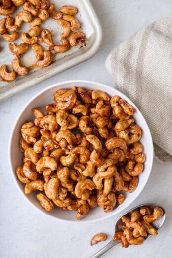 Honey roasted cashews on a small bowl with a spoonful nearby and baking sheet of more snacking nuts nearby.