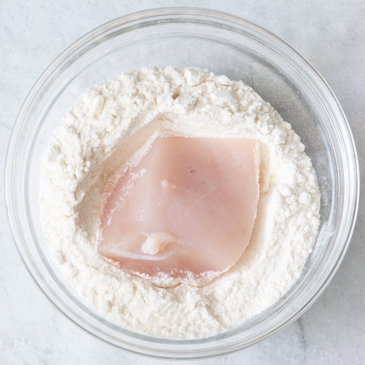 Small piece of uncooked chicken breast on top of a bowl of flour to be dredged.