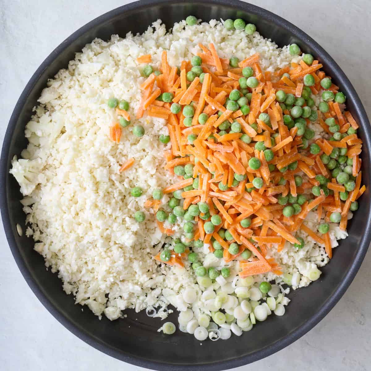 Cauliflower rice in large skillet with white parts of green onion, peas and carrots.