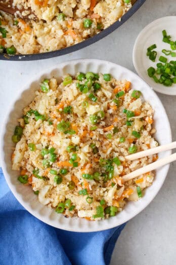 Serving of riced cauliflower fried rice in a bowl with chopsticks dipped inside with skillet nearby.