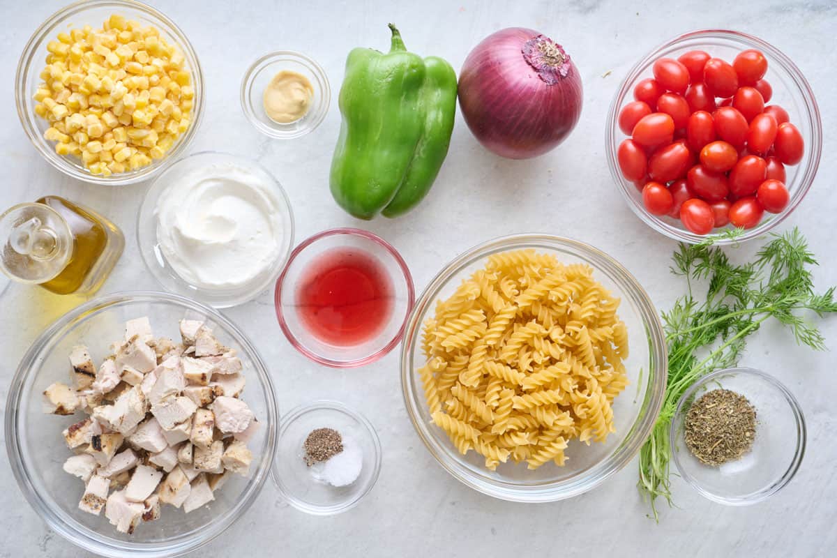 Ingredients for recipe before prepping: corn, oil, diced cooked chicken, greek yogurt, dijon mustard, red wine vinegar, salt and pepper, bell pepper, red onion, fusilli pasta, grape tomatoes, fresh dill, and Italian seasoning.