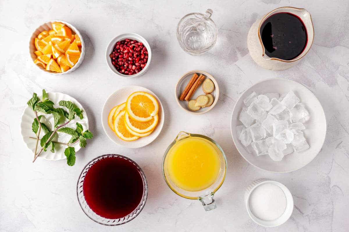 Ingredients for recipe in individual dishes: orange pieces and slices, fresh mint, pomegranate arils, cinnamon and sliced ginger, water, pomegranate juice, cranberry juice, orange juice, ice and sugar.