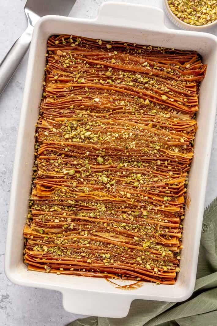 Crinkle cake in baking dish garnished with crushed pistachio and a small dish of more pistachios and serving spatula nearby.